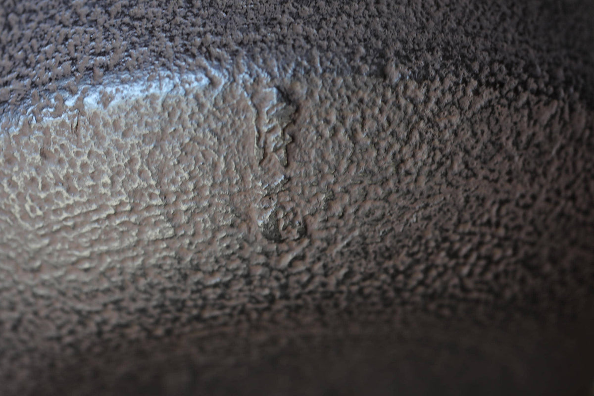 a close-up view of a textured surface, possibly a metal or concrete material, with a rough, uneven texture and a small hole or puncture mark visible.
