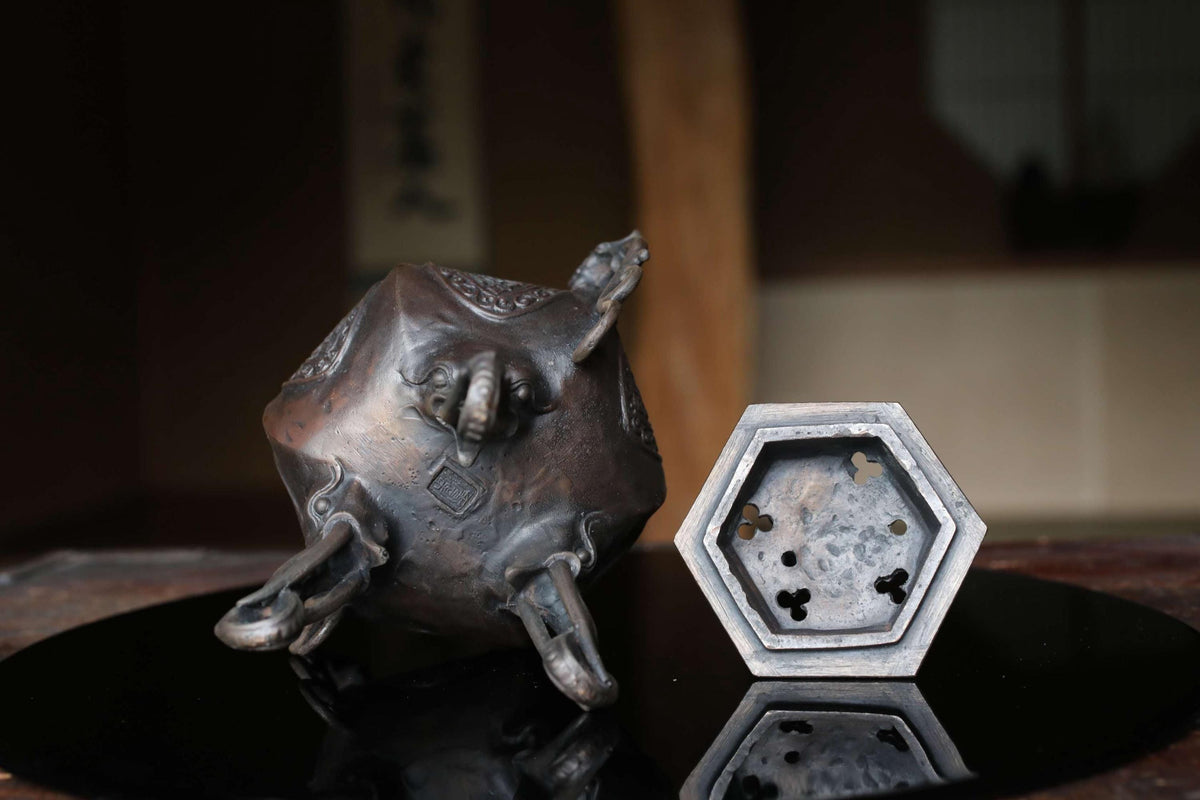 a bronze incense burner with a hexagonal base, placed on a reflective surface.