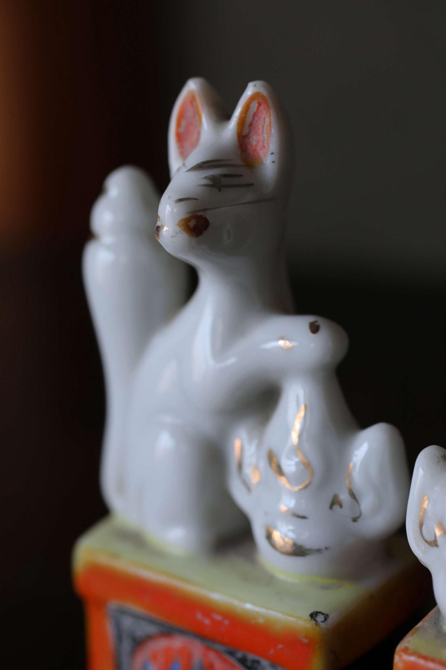 A close-up image of a white ceramic figurine, possibly a cat or rabbit, sitting on a small orange and yellow base.