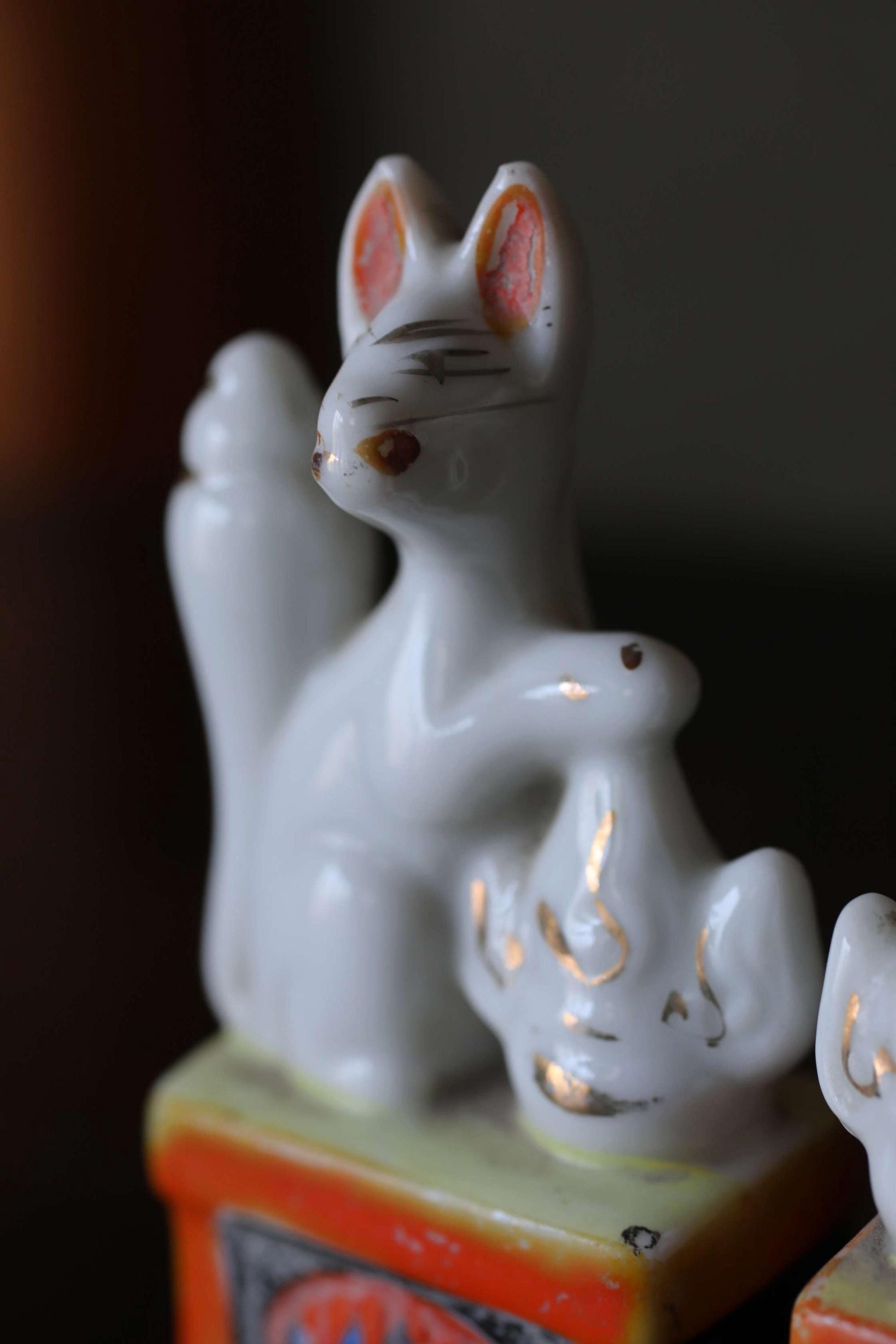 A close-up image of a white ceramic figurine, possibly a cat or rabbit, sitting on a small orange and yellow base.