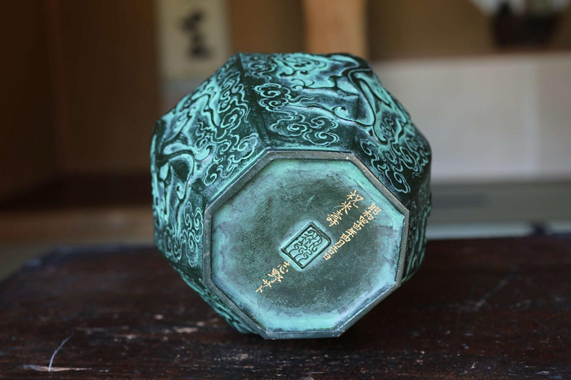 a green, octagonal vase with intricate designs on its surface, sitting on a wooden surface.
