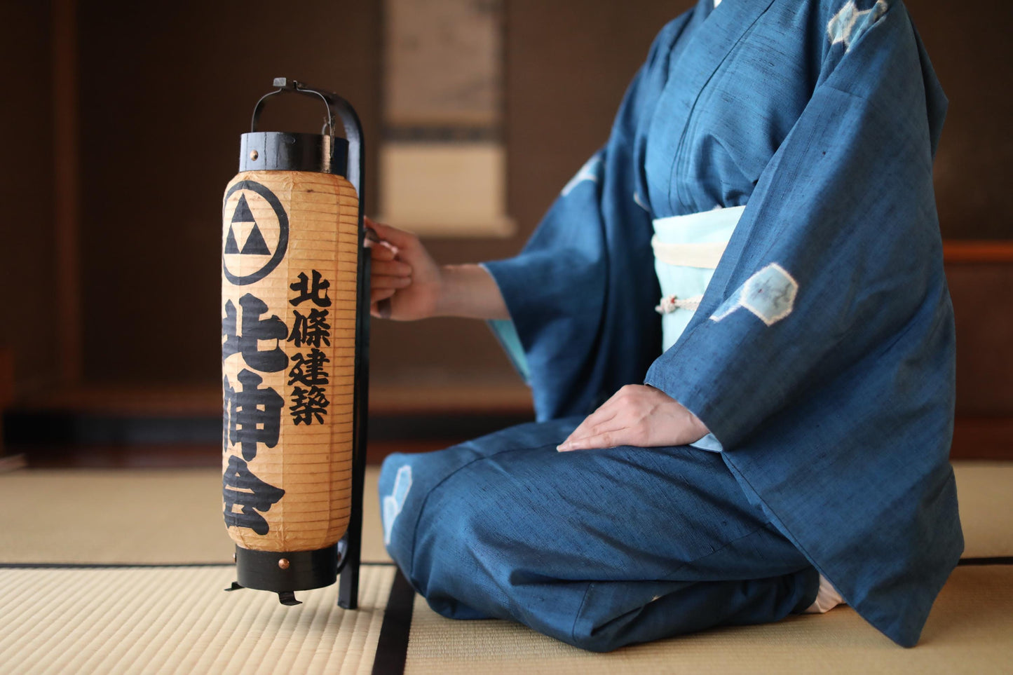 A person wearing a traditional Japanese kimono is seated on the floor, holding a wooden lantern with Japanese characters on it.
