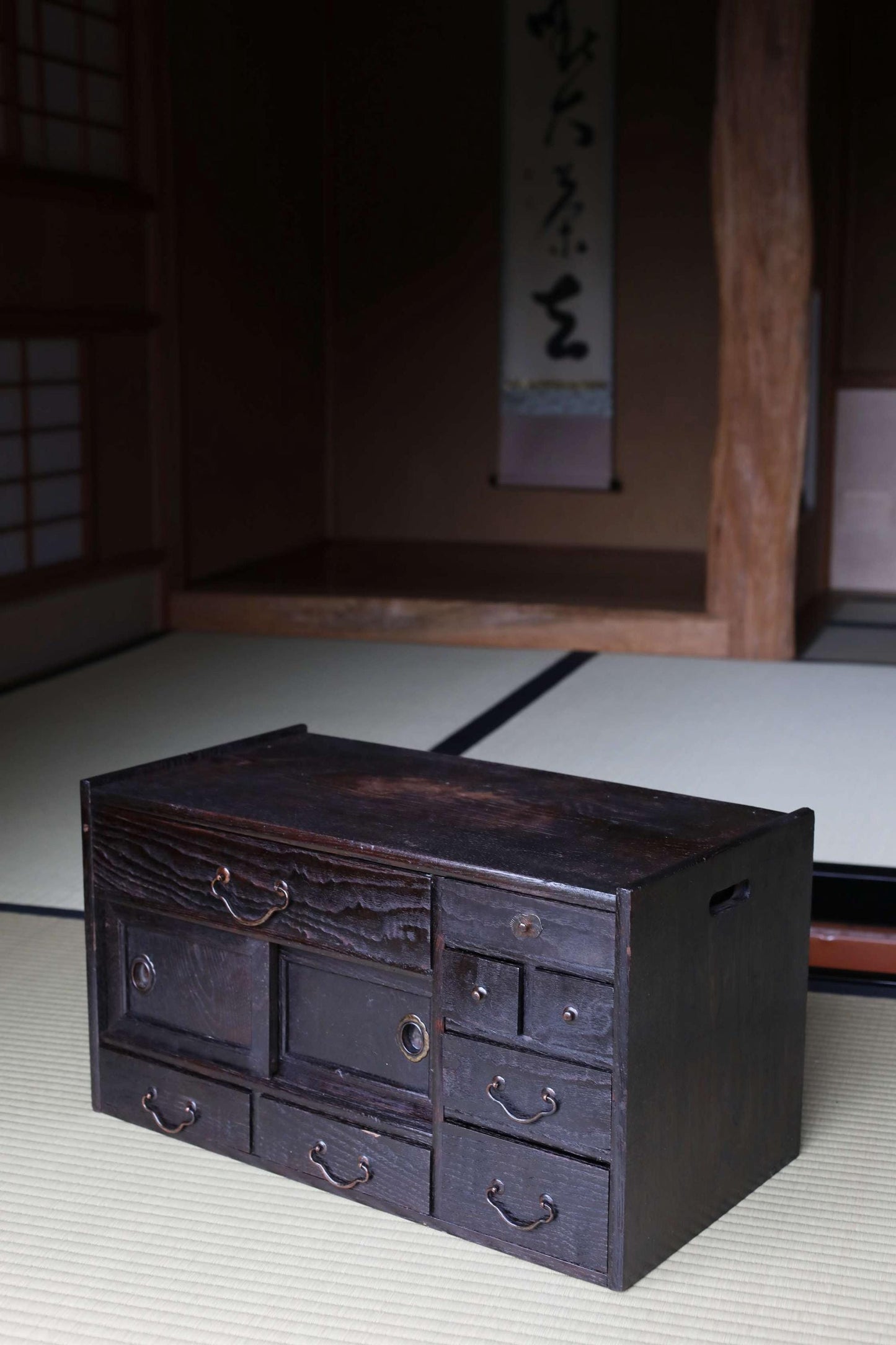 A dark wooden cabinet with multiple drawers sits on a tatami mat in a dimly lit room with a wooden pillar and a Japanese-style banner hanging on the wall.