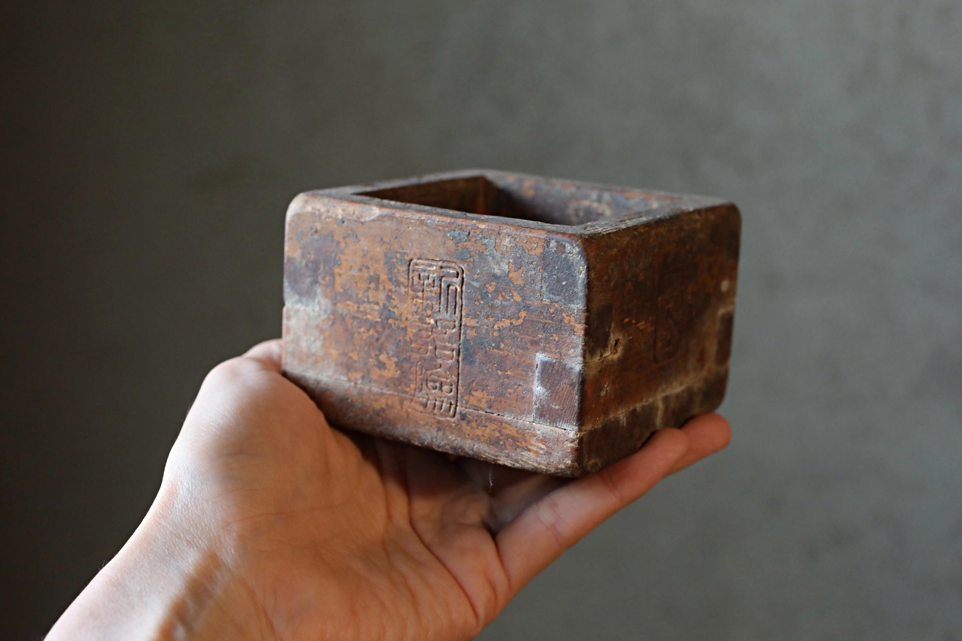 A hand is holding a rusted metal box with a carved design on it.