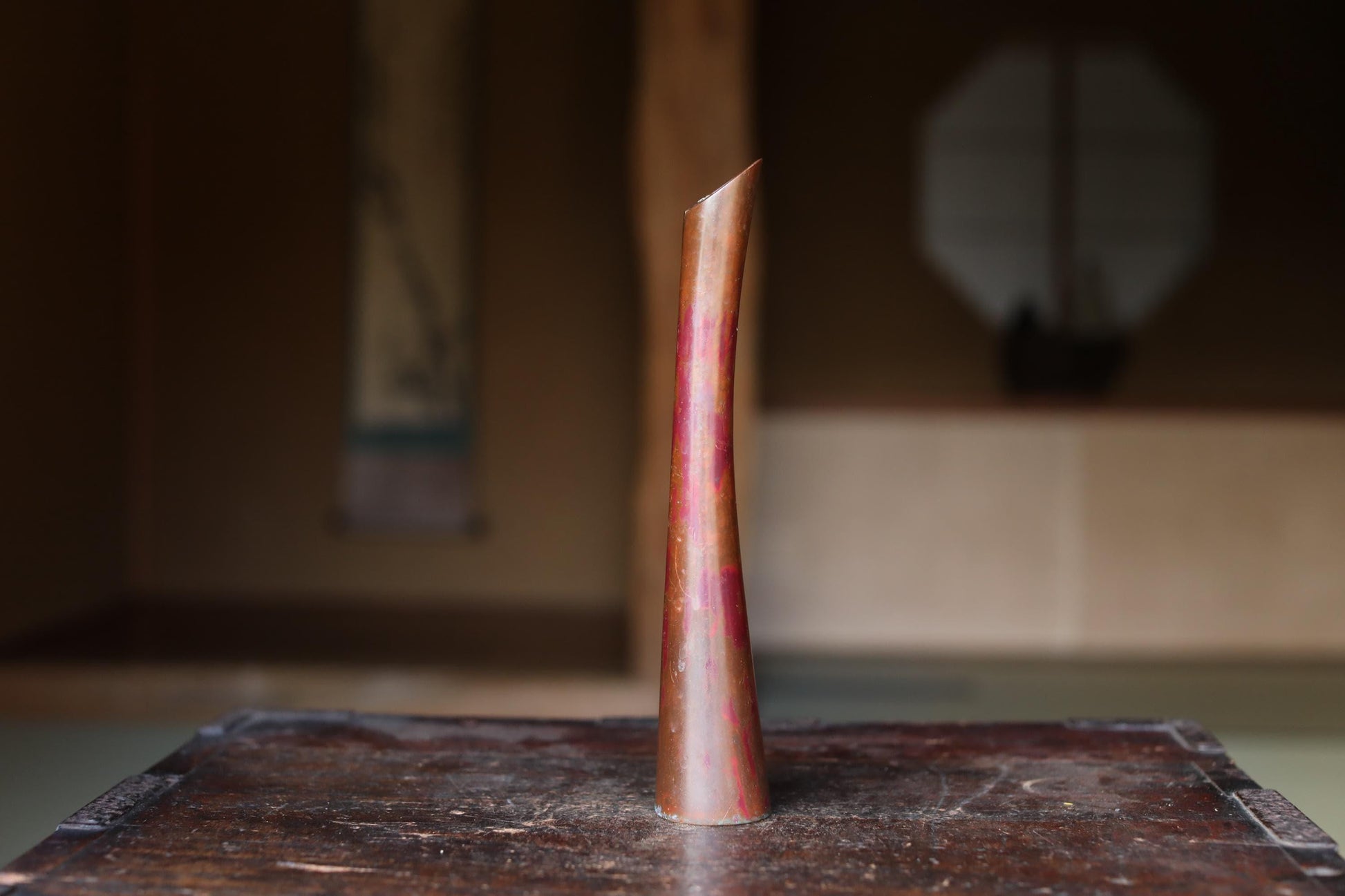 A tall, narrow copper vase with a reddish-brown hue sits on a wooden surface, with a blurred background suggesting an indoor setting.