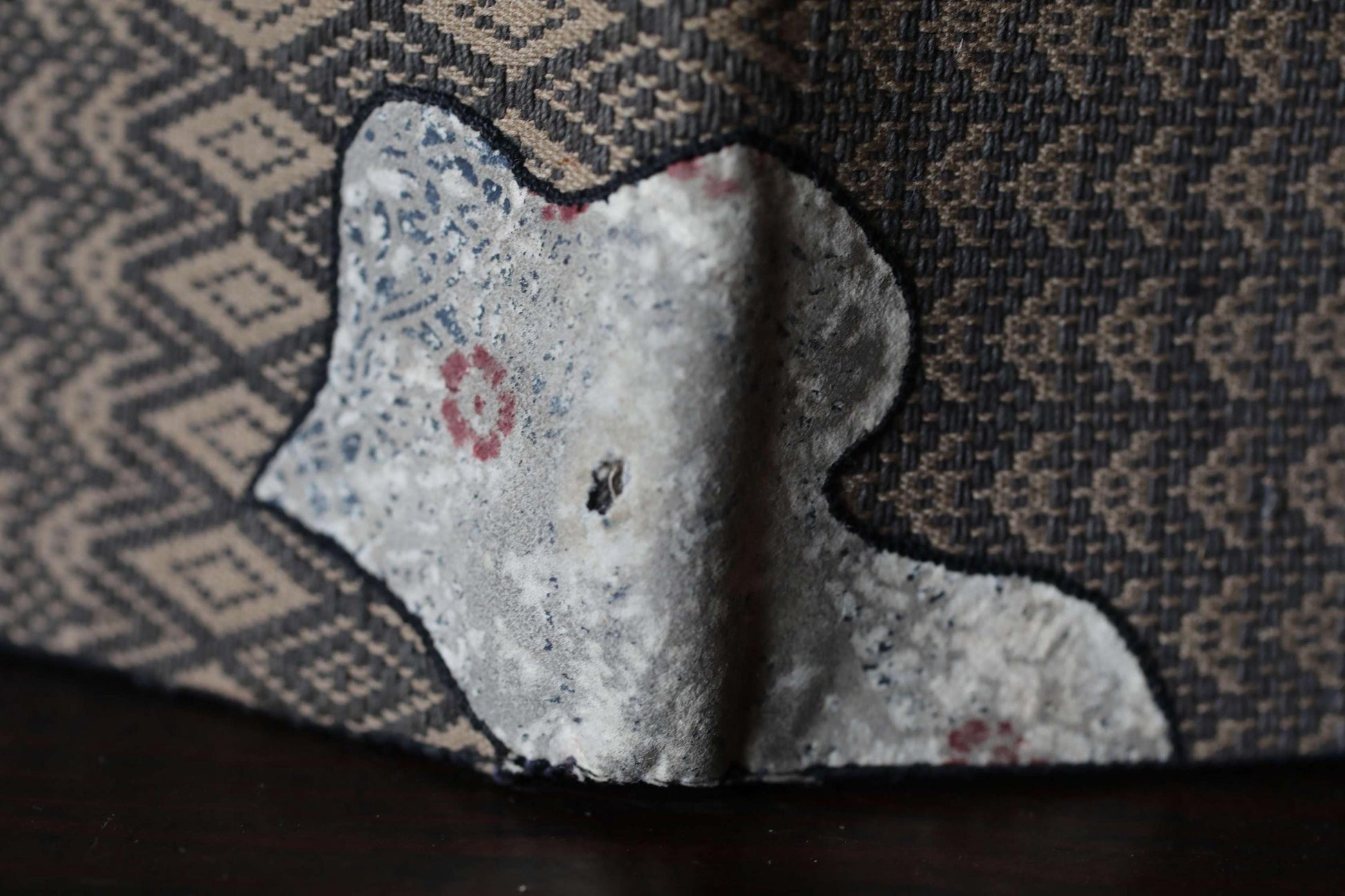 a close-up view of a damaged or worn fabric, possibly a piece of clothing or a textile, with a hole or tear in the center. The fabric appears to be a dark-colored material with a patterned design.