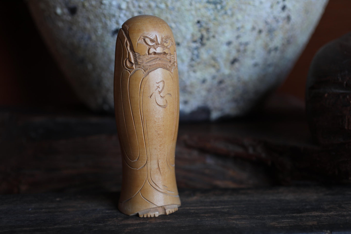 A wooden figurine of a deity or spiritual figure is displayed on a wooden surface, with a large ceramic bowl or vase in the background.