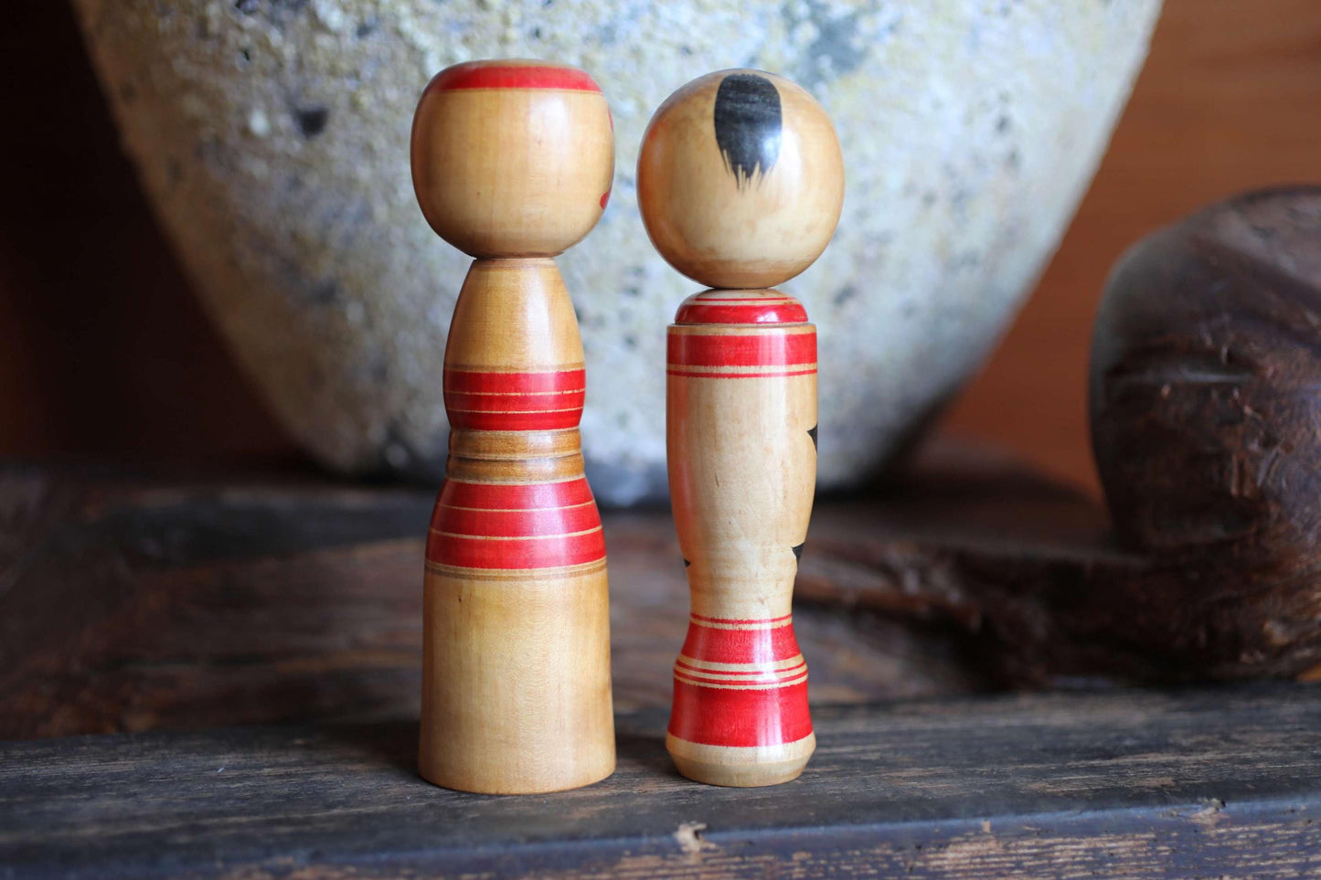 Two wooden figurines with red and brown accents are displayed on a wooden surface, with a large white ceramic object in the background.