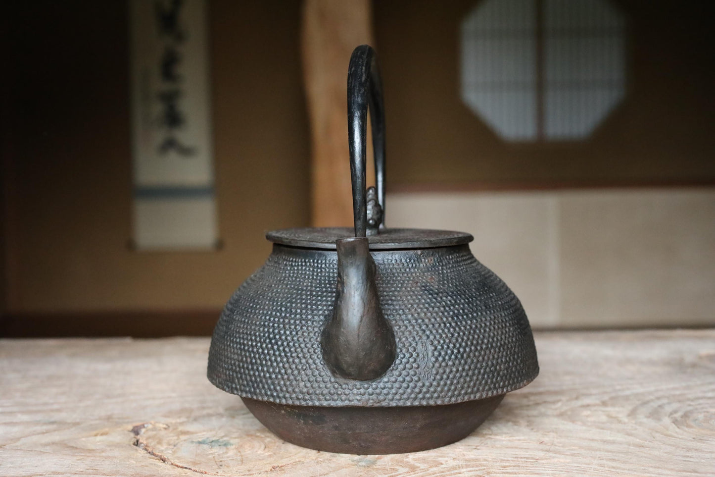 An antique, black metal teapot with a curved handle and spout sits on a wooden surface.