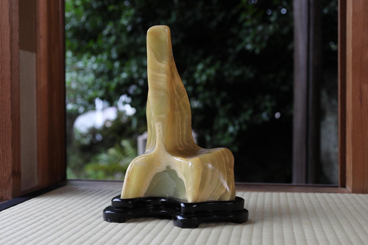 A yellow, abstract sculpture is displayed on a black stand in front of a window with a view of trees.