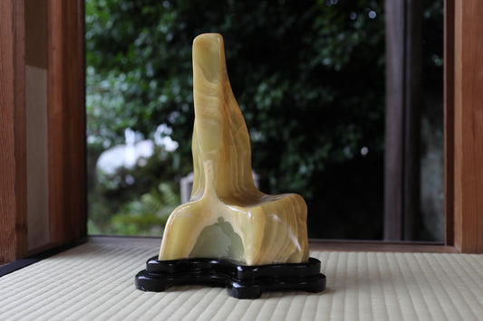 A yellow, abstract sculpture is displayed on a black stand in front of a window with a view of trees.