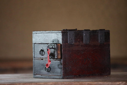 An old, wooden chest with metal clasps and a red ribbon tied around the handle, sitting on a wooden surface.