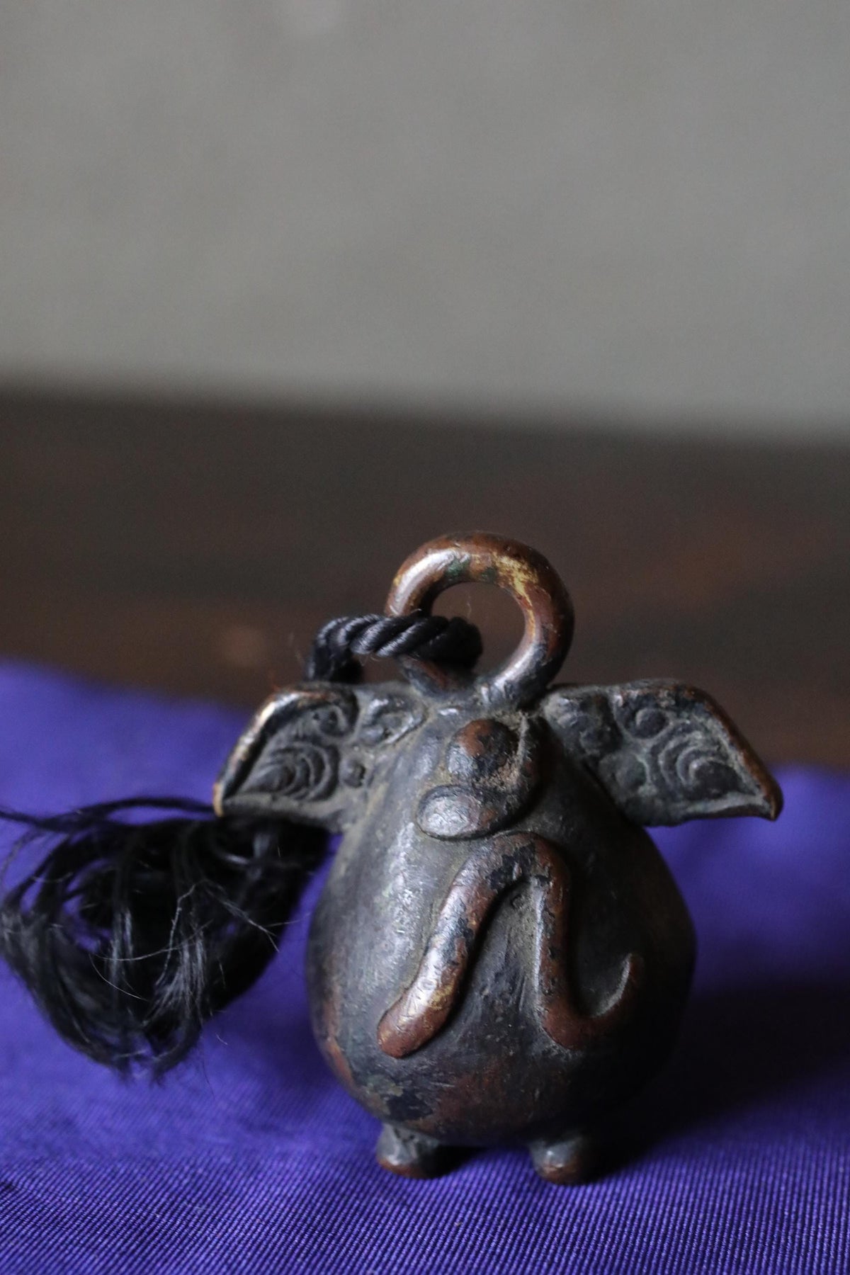 A small, intricately designed metal bell with a curved handle and a tassel hanging from its neck, resting on a purple surface.