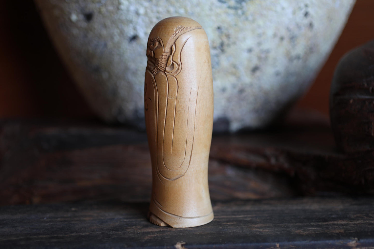 A wooden figurine with an intricate design sits on a wooden surface, with a large ceramic bowl visible in the background.