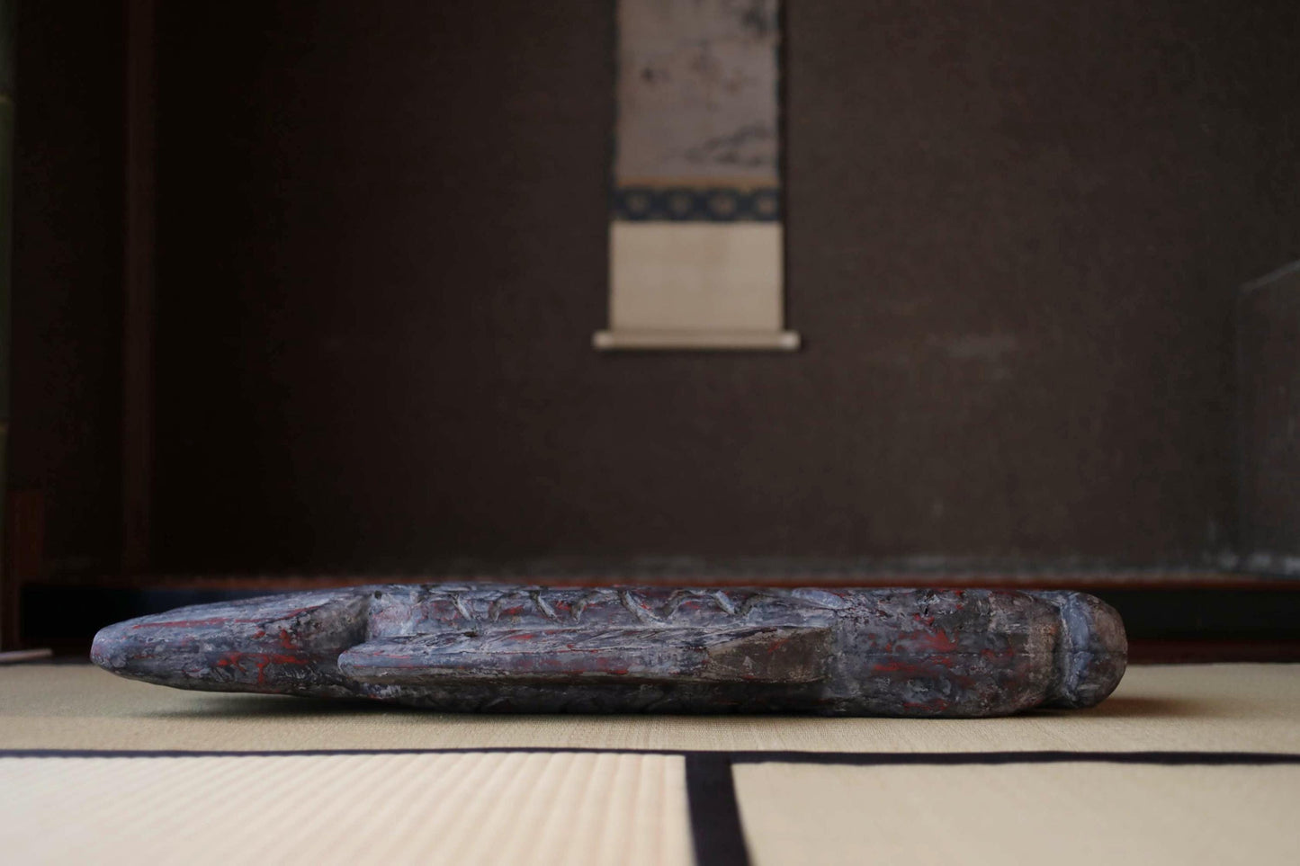 a wooden object, possibly a sculpture or a piece of furniture, resting on a tatami mat in a room with a dark background.