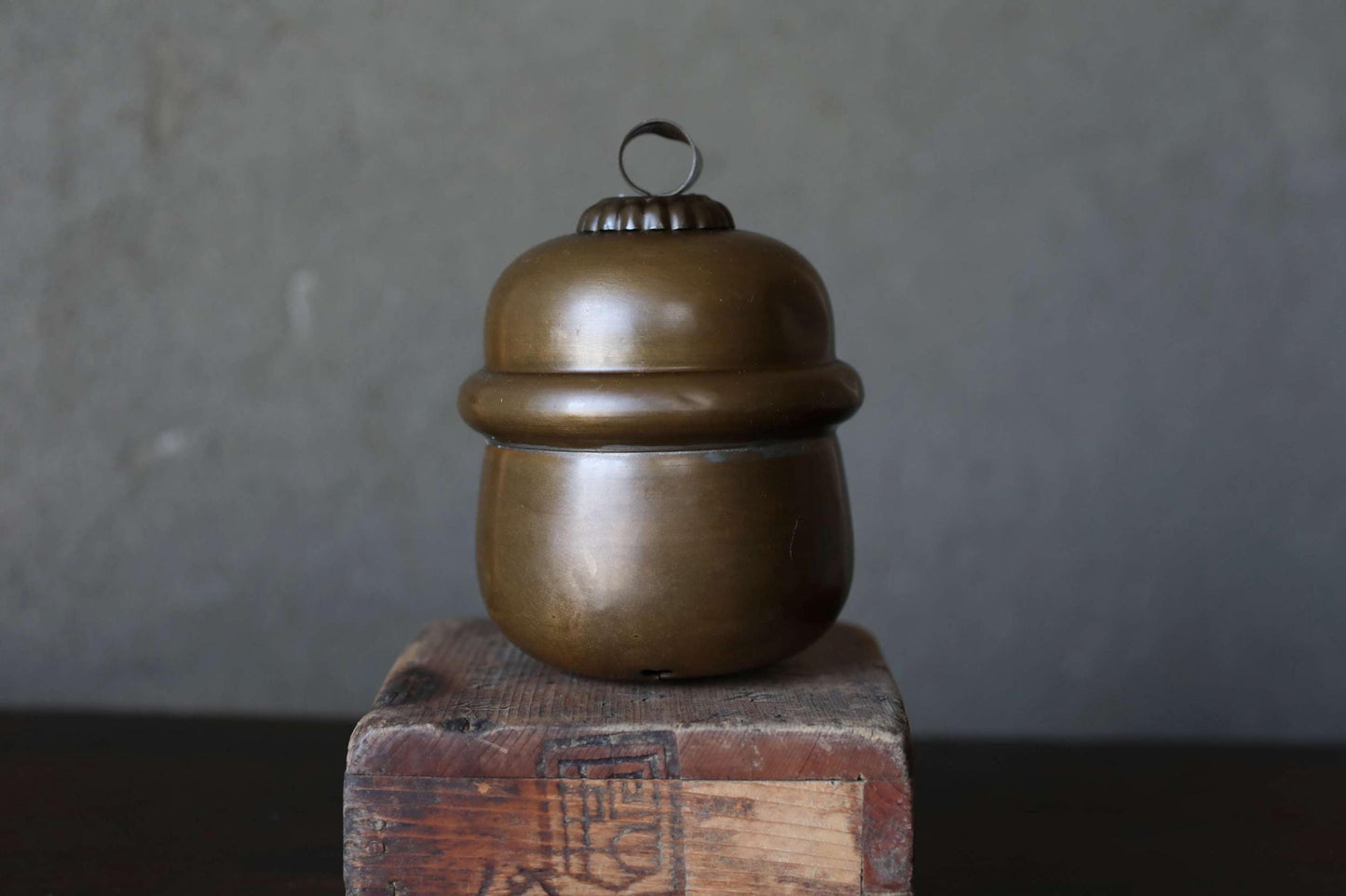 A brass container with a round lid sits on a wooden block against a gray background.