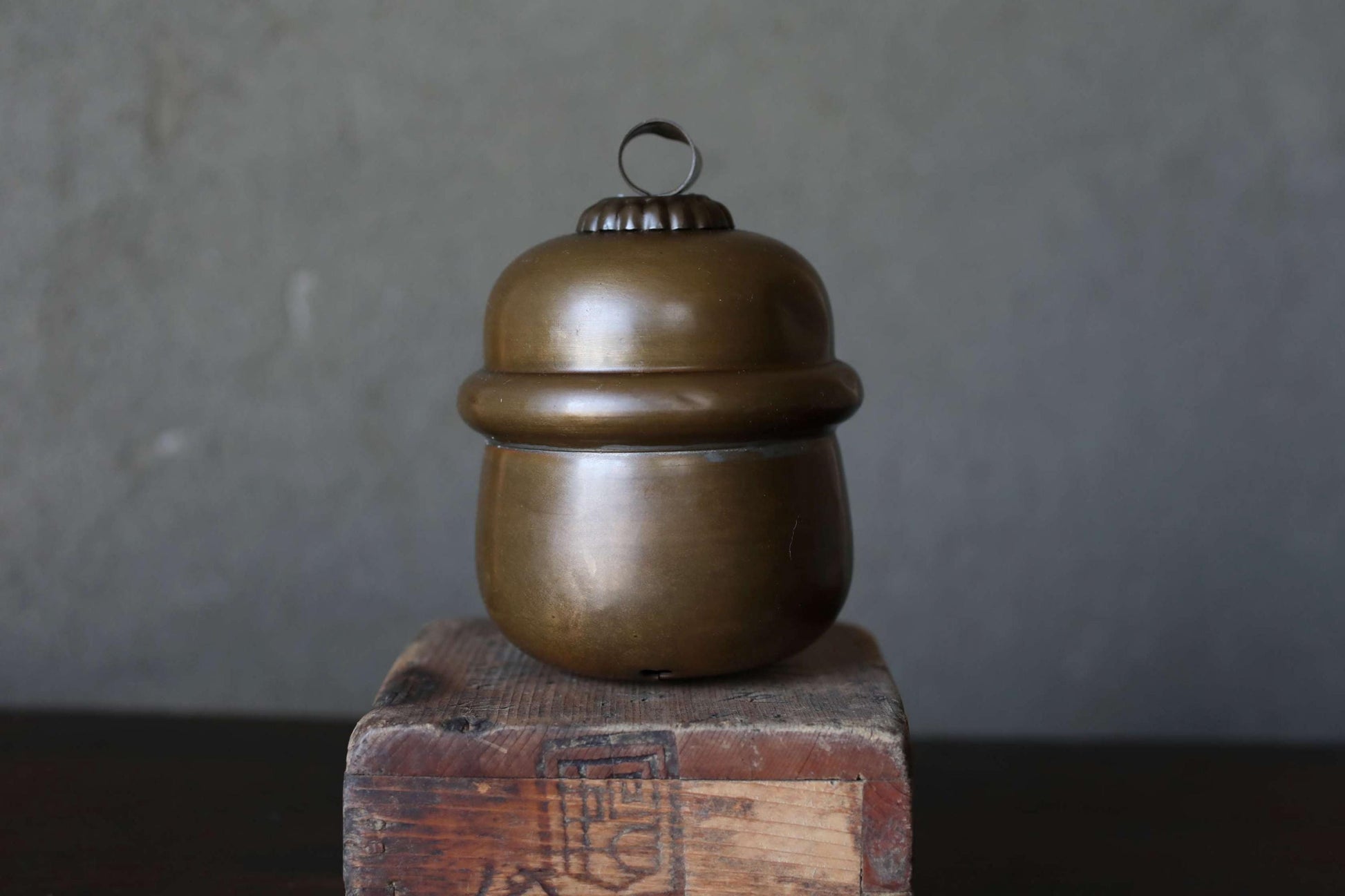 A brass container with a round lid sits on a wooden block against a gray background.