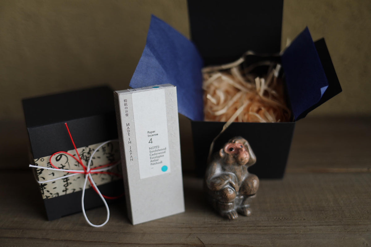 a small ceramic monkey figurine, a black box with a white label, and a small gift box with a red ribbon tied around it, all placed on a wooden surface.