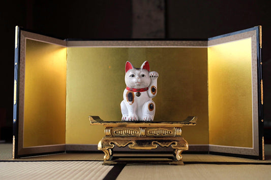 A white ceramic cat figurine with red ears is sitting on a decorative golden pedestal, placed on a tatami mat.