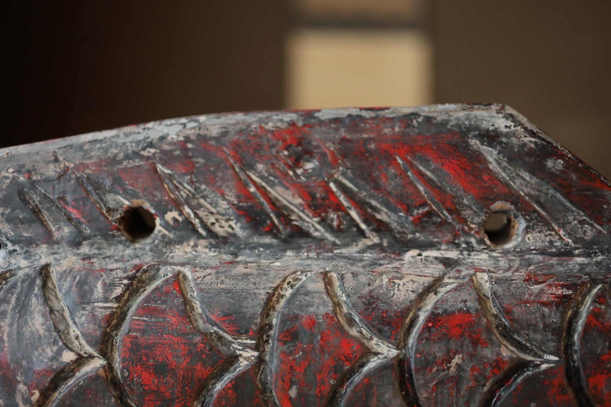a close-up view of a weathered, textured surface with a red and black color scheme. The surface appears to be a wooden object, possibly a piece of furniture or a decorative item. The surface has a rough, uneven texture, and there are two holes visible on the surface.