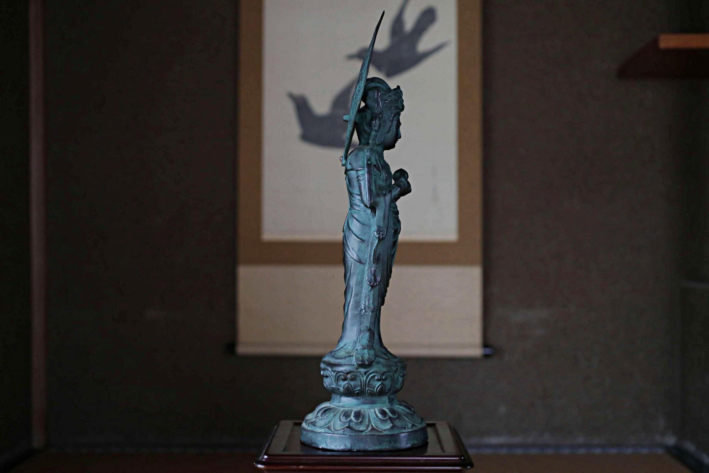 A bronze statue of a deity stands on a wooden base, with a shadow cast on the wall behind it.