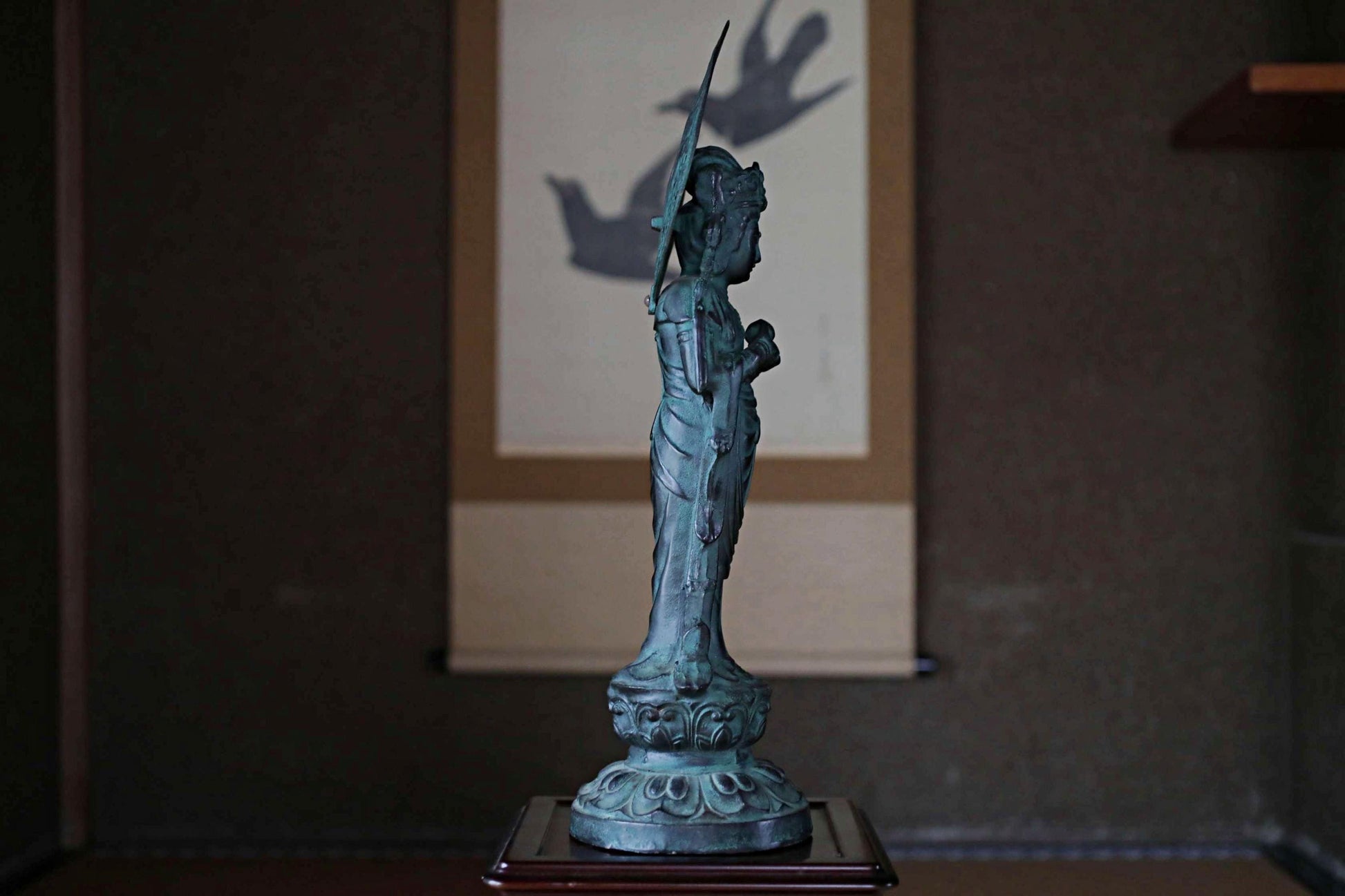 A bronze statue of a deity stands on a wooden base, with a shadow cast on the wall behind it.