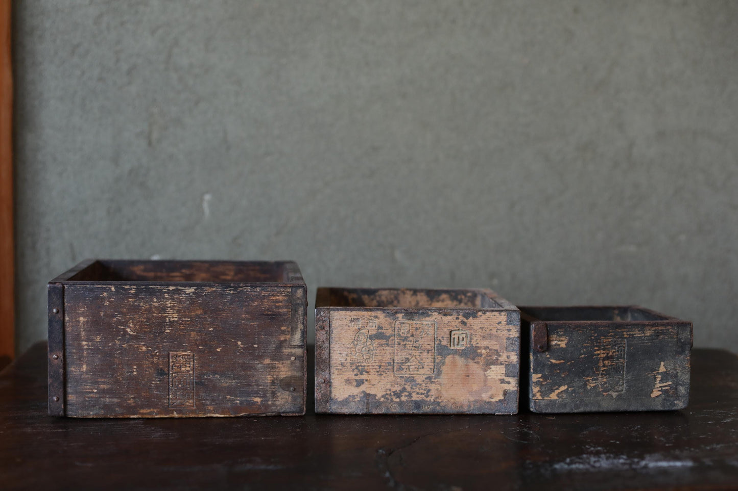 Three old, weathered wooden crates or boxes are arranged on a dark surface, with a textured gray wall in the background.