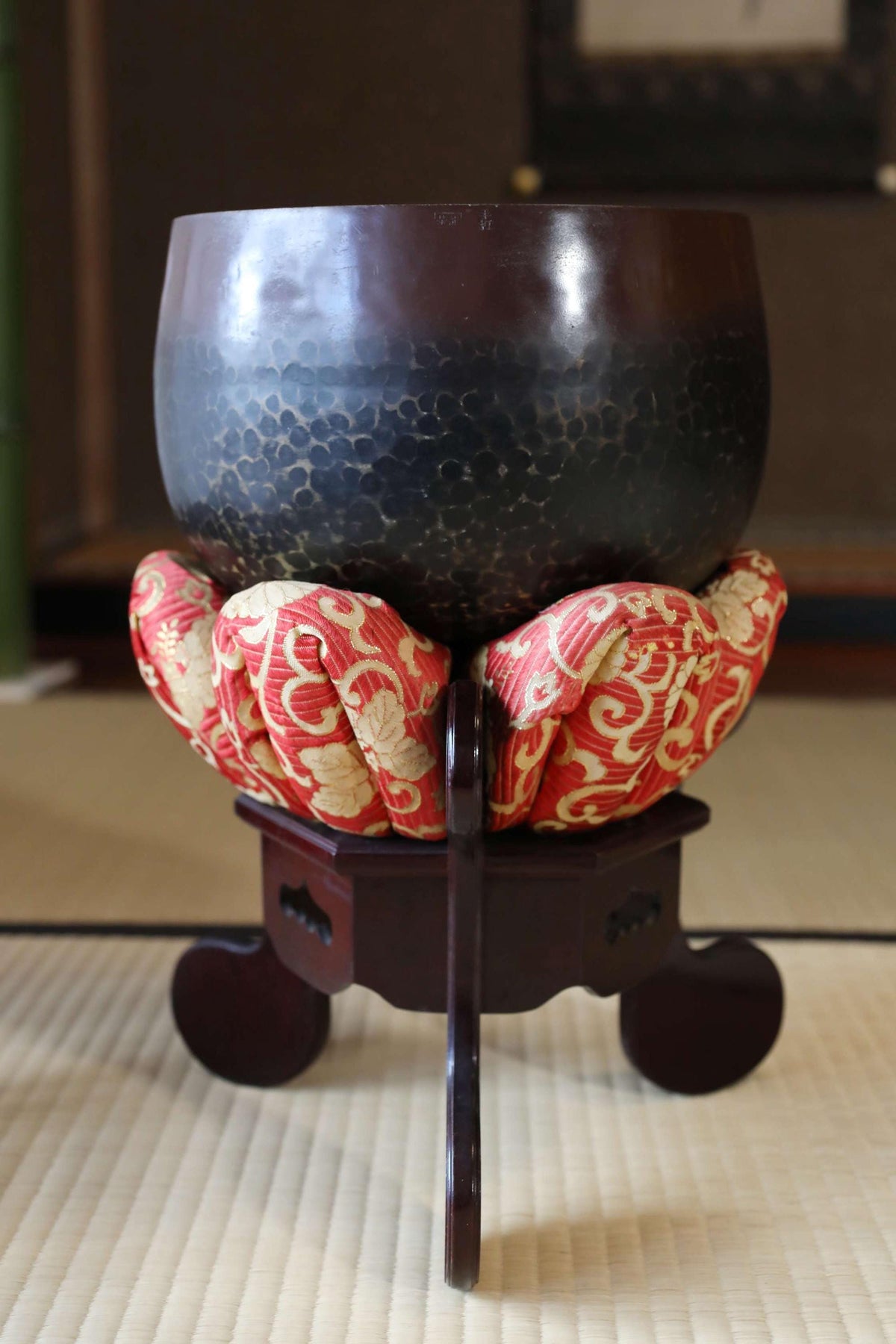 A large, dark bowl sits on a wooden stand with red and gold fabric accents, placed on a tatami mat.