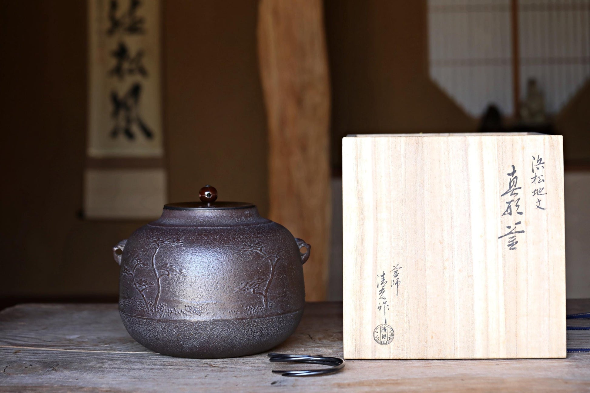 以下が含まれることがあります： 木製の蓋と装飾的なデザインが施された、濃い灰色の鋳鉄製の急須。急須は、日本の文字が書かれた木製の箱の隣にある木製のテーブルの上に置かれています。