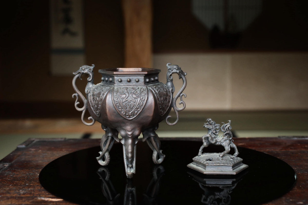 a decorative metal urn with an elephant-like design on its front, placed on a reflective surface. Next to the urn is a small metal figurine of a lion.