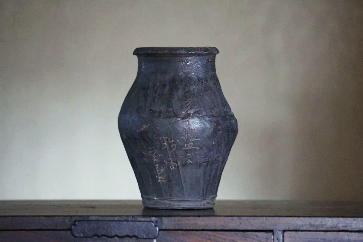 A dark-colored vase with intricate designs and patterns, sitting on a wooden surface against a plain background.
