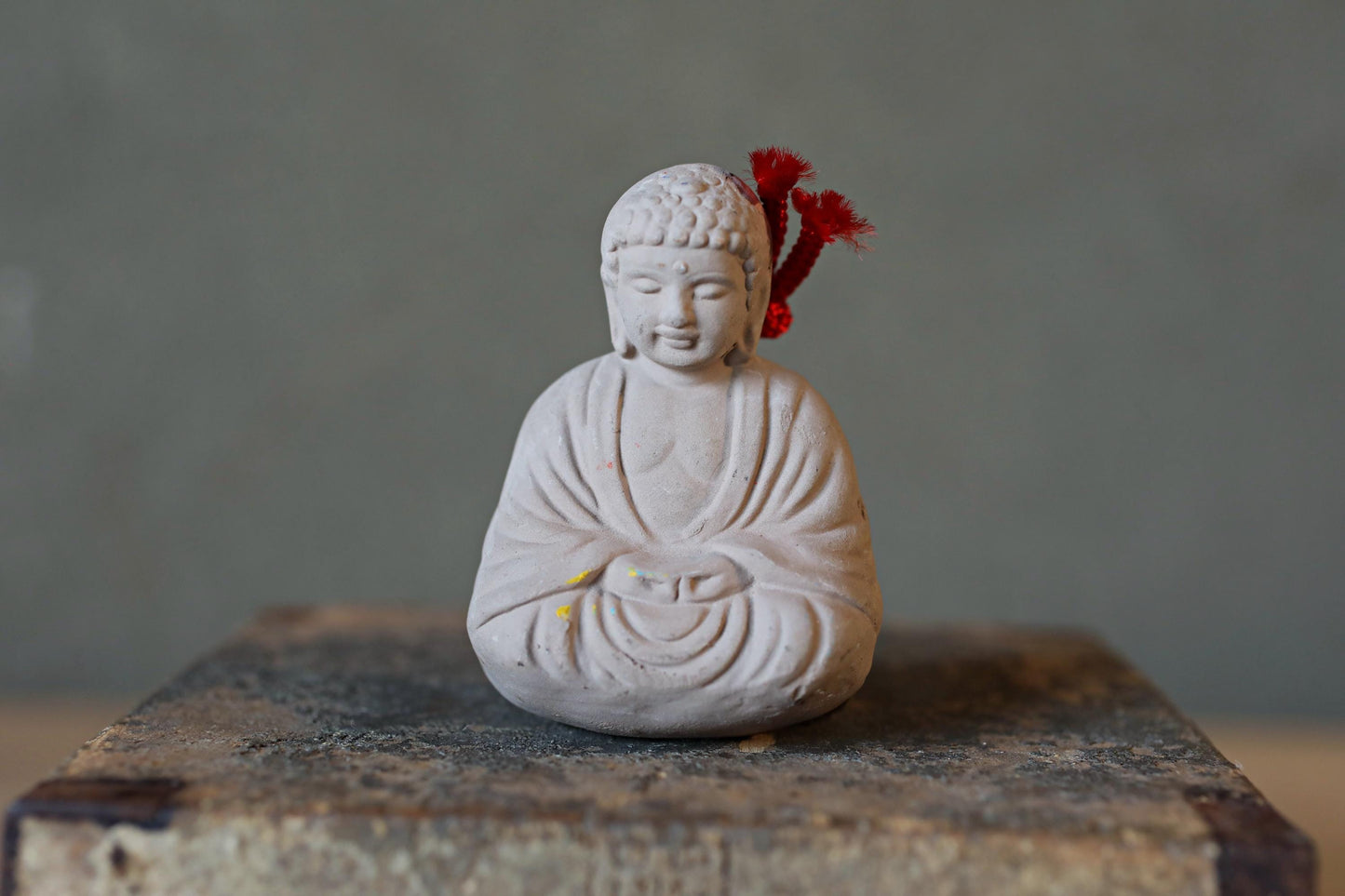 A small white Buddha statue with a red tassel on its head, sitting on a wooden surface against a gray background.