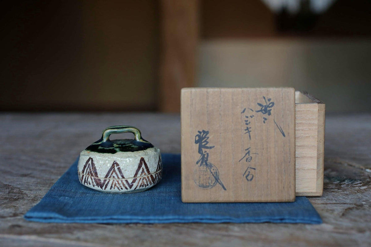 a small ceramic container with a handle, placed on a blue cloth on a wooden surface. Next to the container is a wooden box with Japanese characters on it.