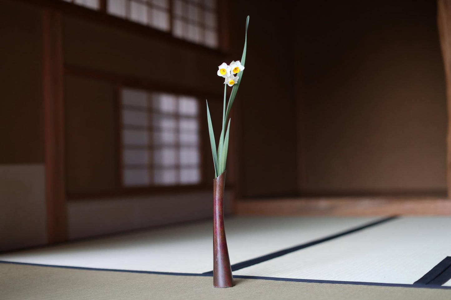 A single white and yellow flower in a brown vase is placed on a tatami mat in a traditional Japanese room.
