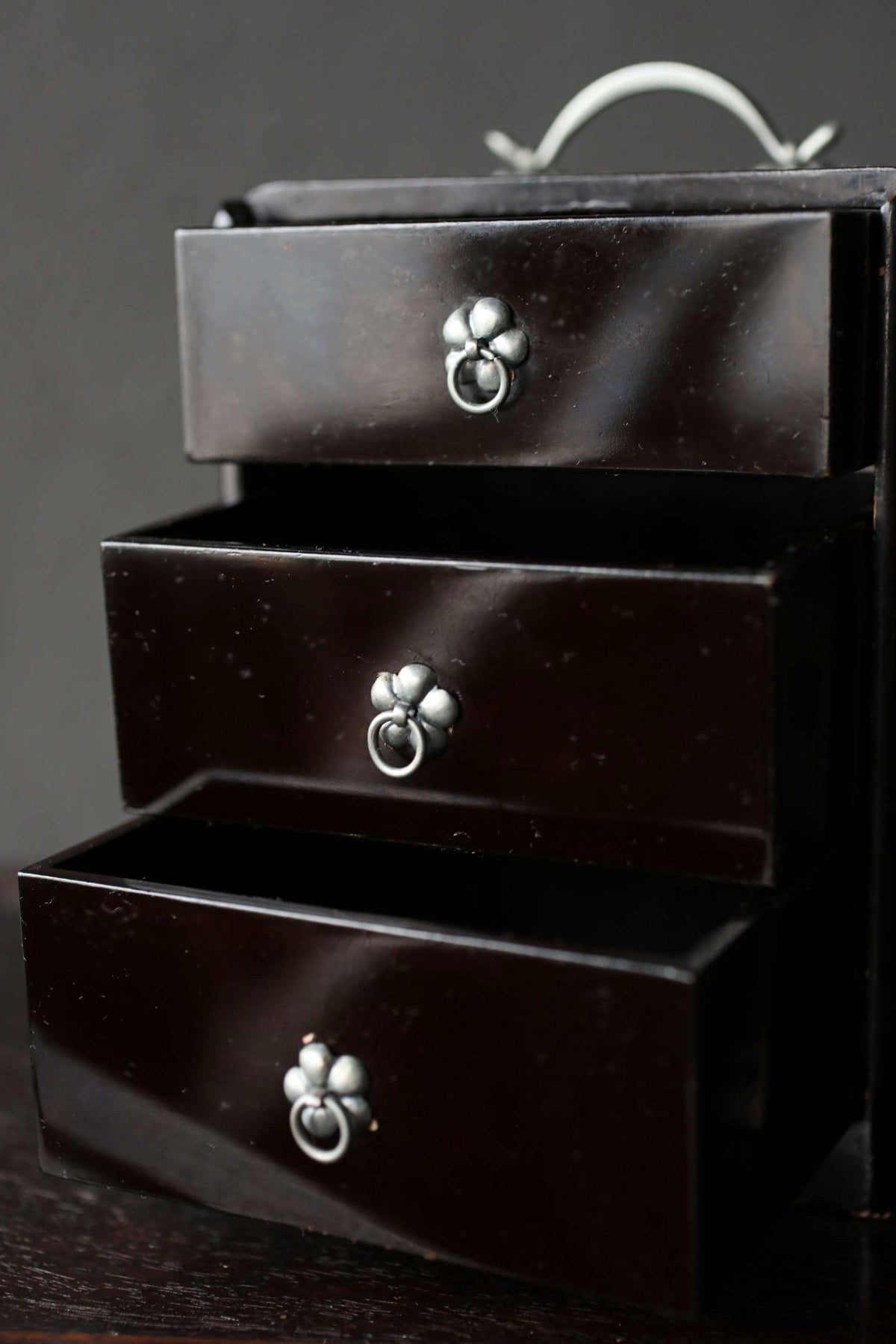 A close-up view of a dark wooden drawer with three metal handles, set against a dark background.