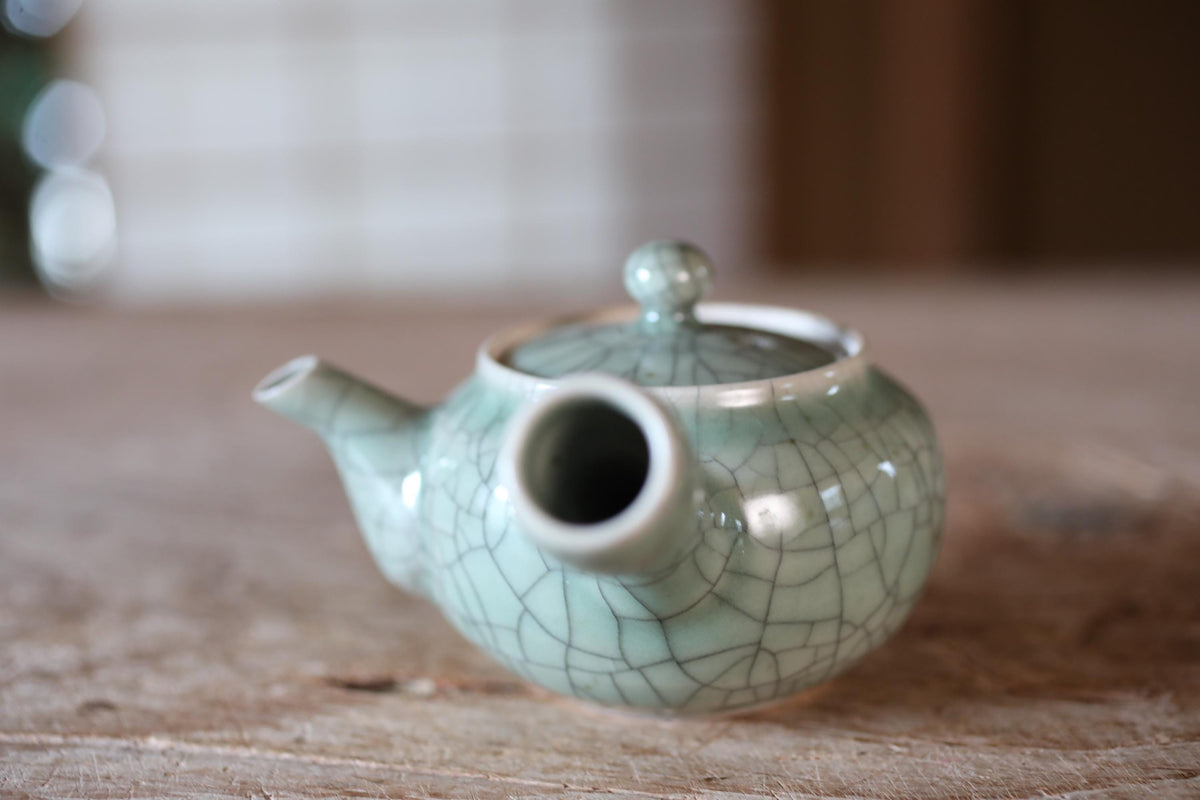 A ceramic teapot with a green and white color scheme, featuring a handle and spout, sits on a wooden surface.