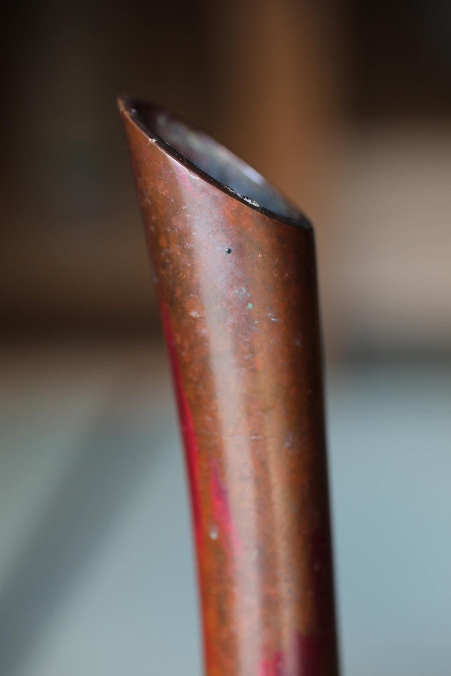 A close-up image of a reddish-brown metal object, possibly a pipe or tube, with a slight curve and a small hole at the top.