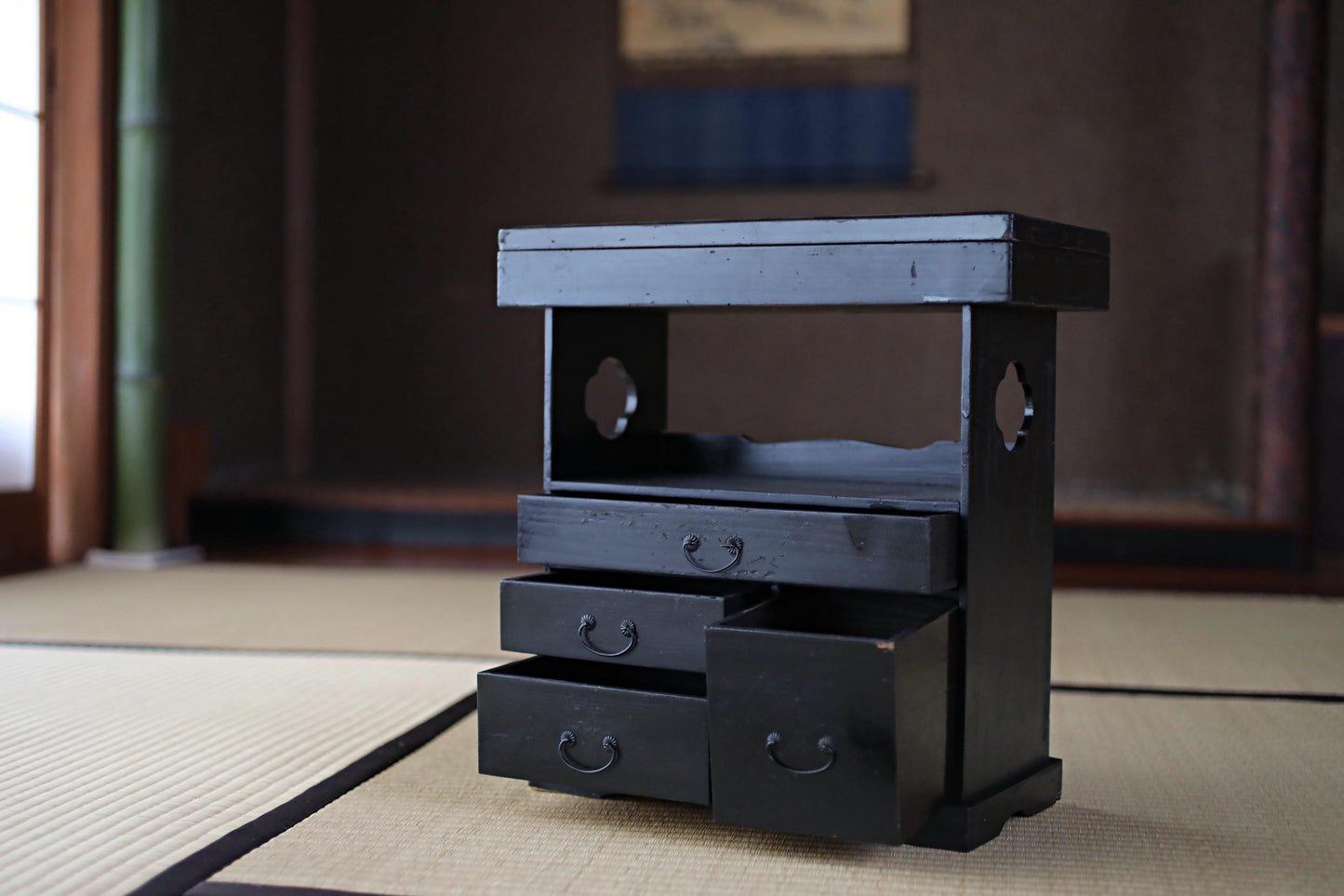 A small black wooden cabinet with three drawers and a shelf, placed on a tatami mat in a room with a window and a bamboo pole.