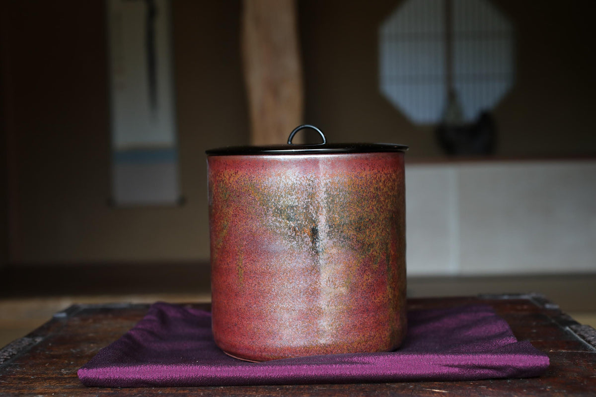 A red ceramic container with a black lid sits on a purple cloth on a wooden surface.