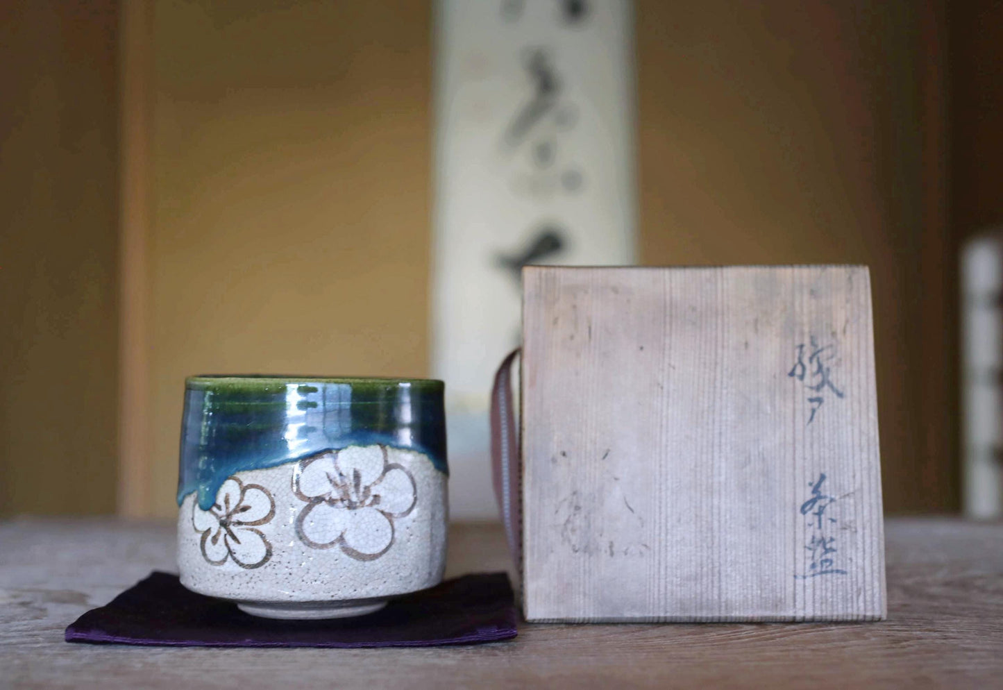 A ceramic cup with a floral design sits on a dark cloth, accompanied by a wooden box with Japanese characters on it, both placed on a wooden surface.