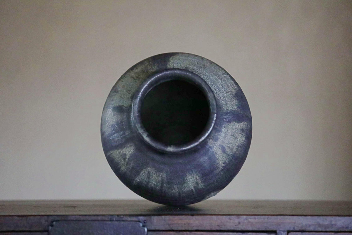 A dark blue ceramic vase with a round shape and a small opening at the top, sitting on a wooden surface against a plain background.