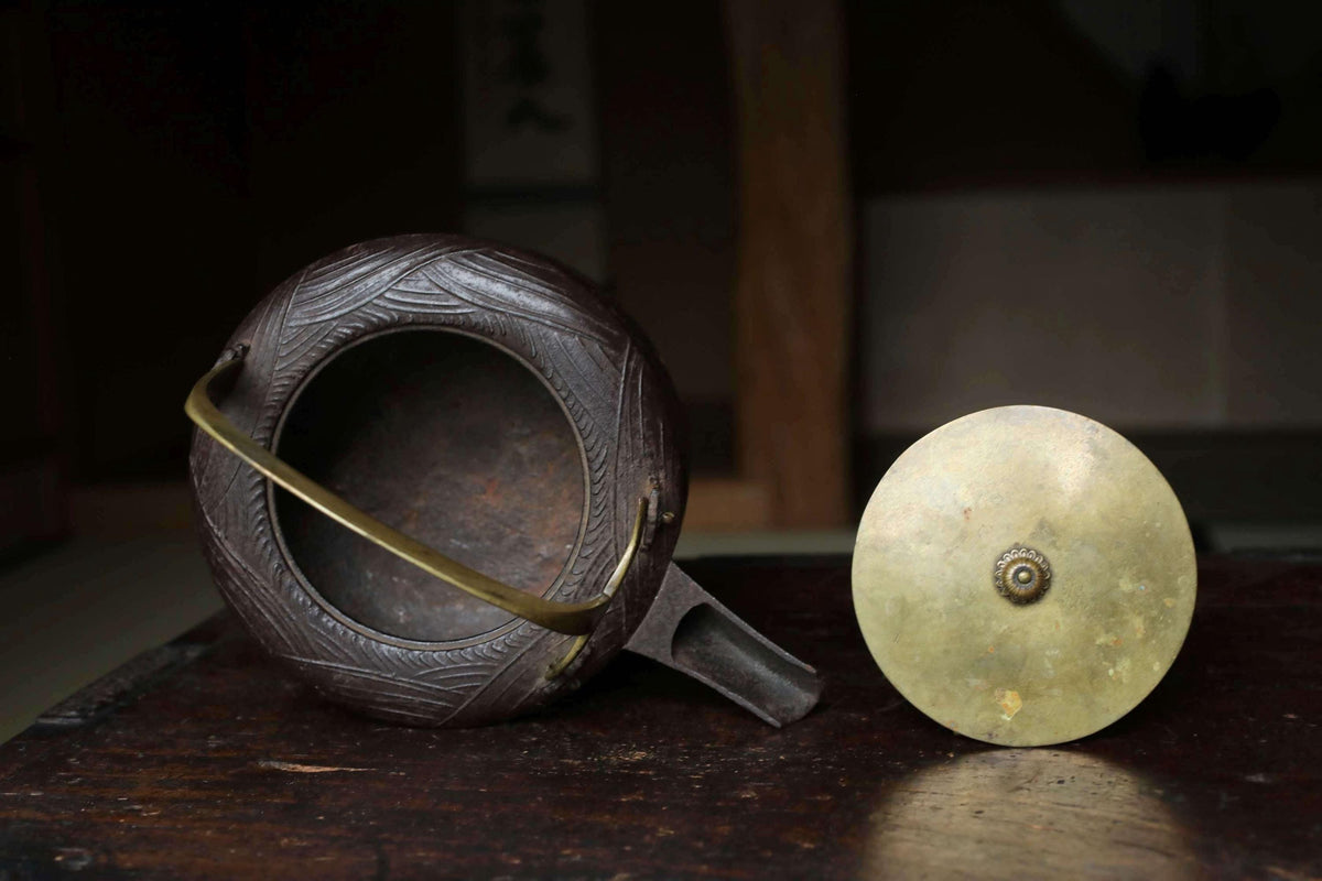 An antique teapot and a brass bell sit on a wooden surface, with the teapot having a dark brown color and intricate designs, and the brass bell having a round shape and a small hole in the center.