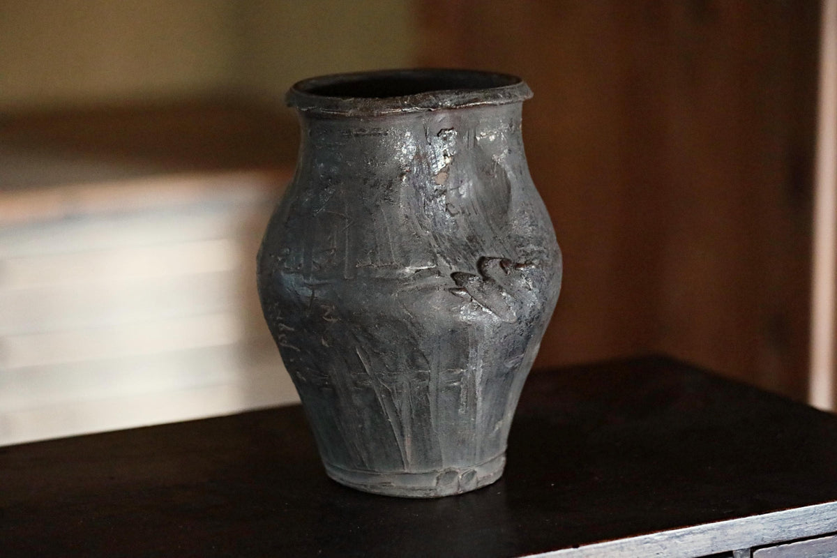 A dark-colored vase with a textured surface sits on a dark surface.