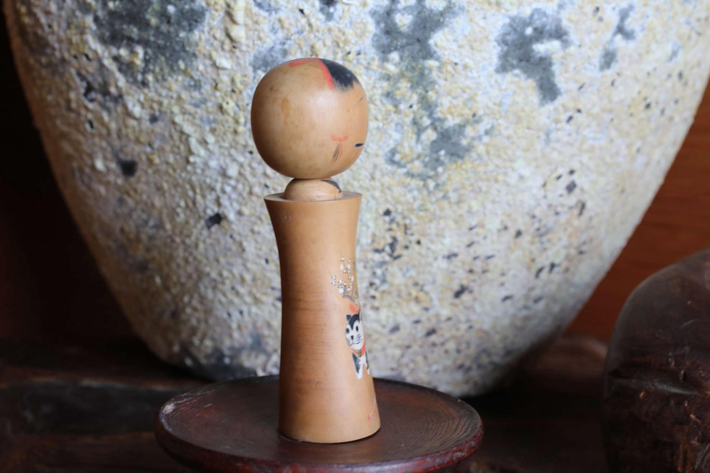 A wooden doll with a carved face stands on a wooden stand in front of a large stone vase.