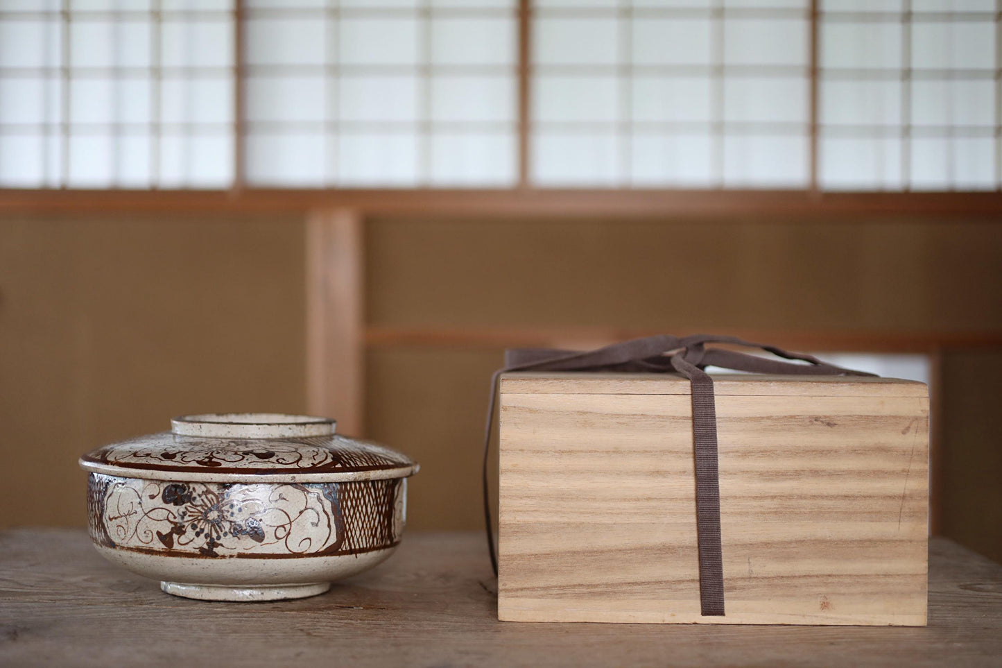 a wooden box with a brown ribbon tied around it, placed next to a ceramic bowl with a floral design on a wooden surface.