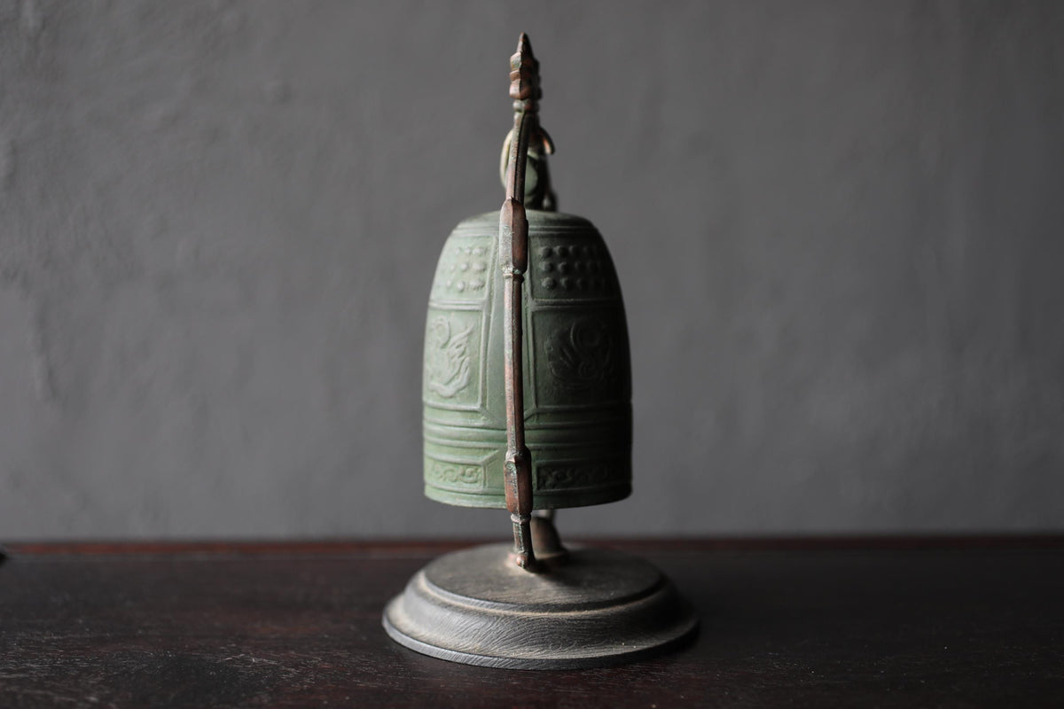 A green metal bell with a decorative design on its surface, placed on a dark surface against a gray background.