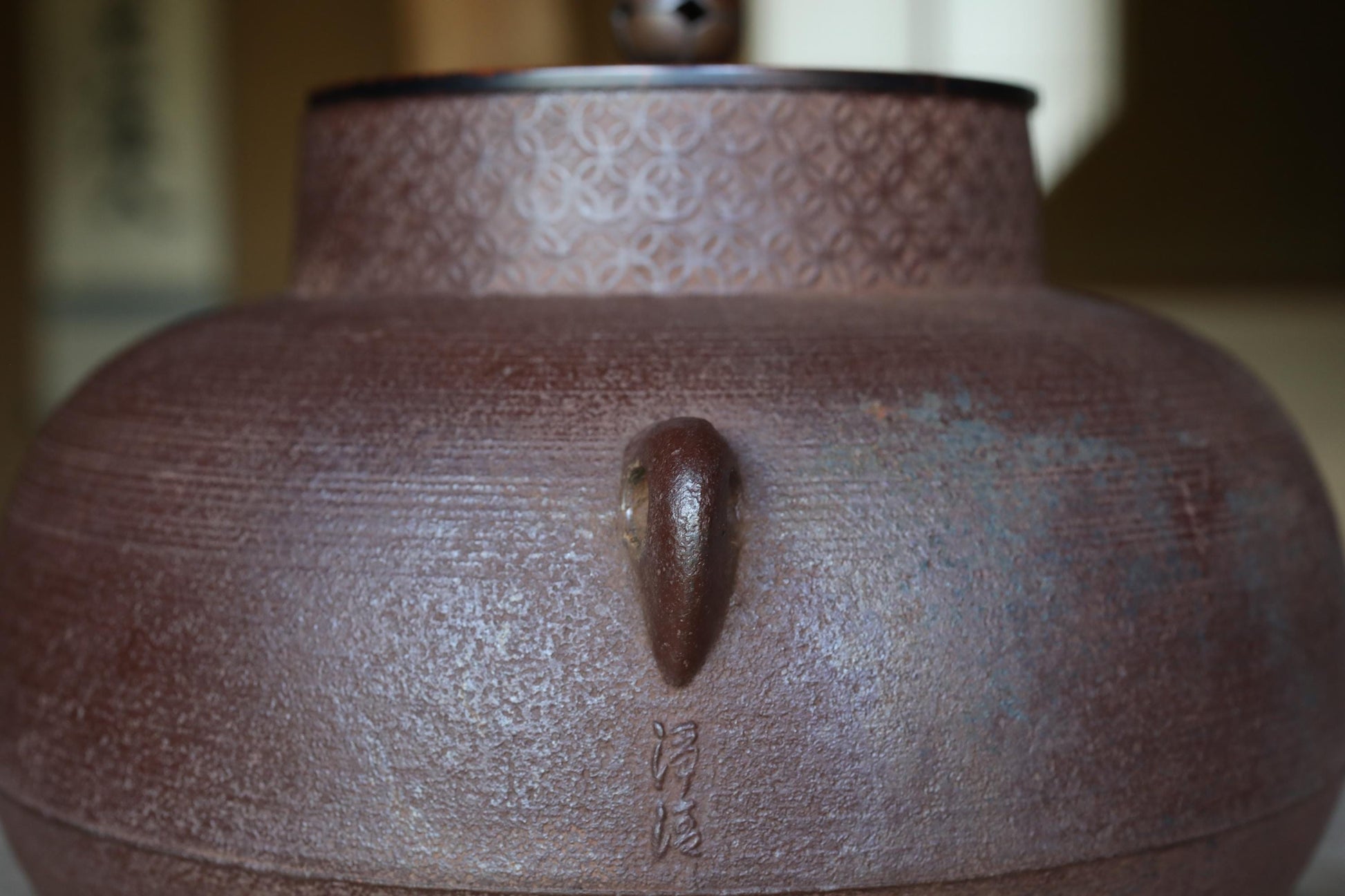 A close-up image of a brown ceramic teapot with a lid and handle, featuring a decorative pattern on the top.