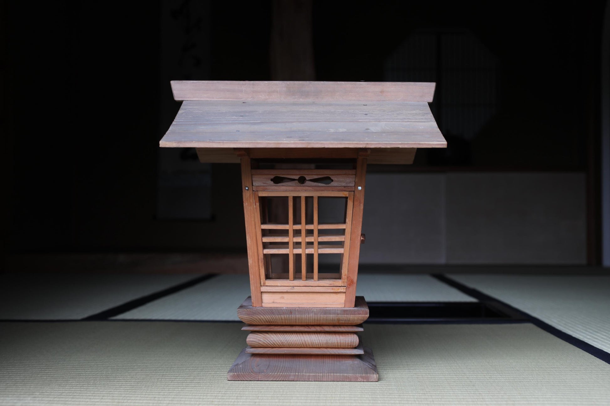 A wooden shrine or altar with a sloping roof and a decorative design, placed on a tatami mat in a dimly lit room.