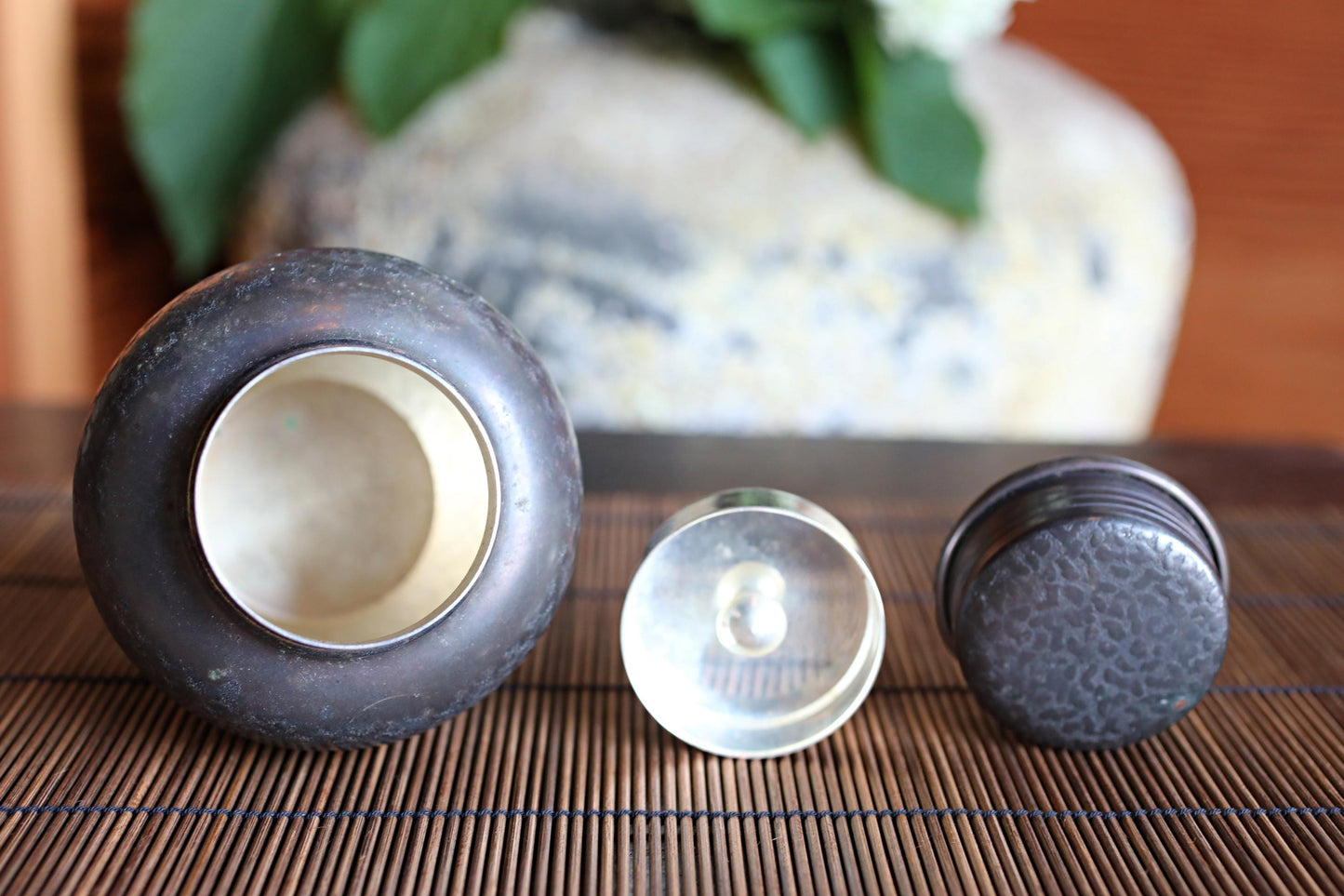 a close-up of three small metal objects, including a round metal container, a small metal lid, and a small metal ring, placed on a bamboo mat.