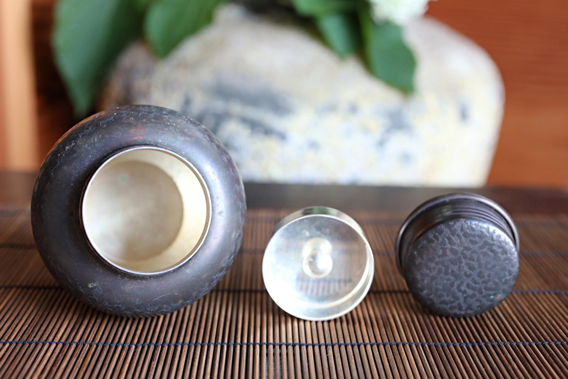 a close-up of three small metal objects, including a round metal container, a small metal lid, and a small metal ring, placed on a bamboo mat.